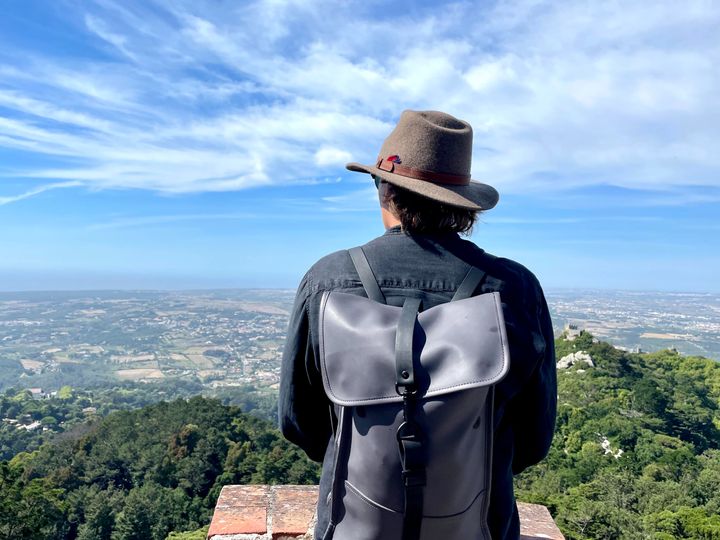 Iz wearing a Rains grey backpack, a black shirt and a hat. Looking over Portugal, Sintra.