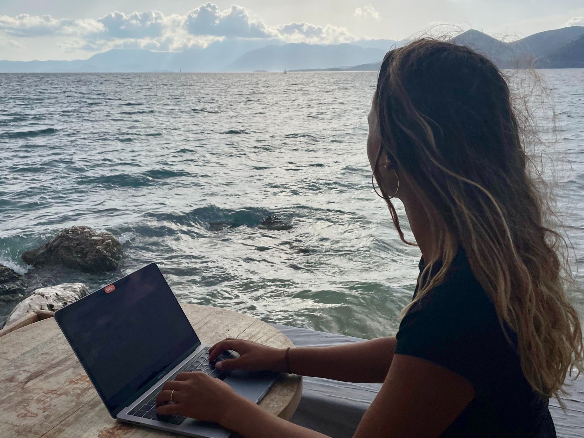 Lau working on her laptop near the ocean. Mountains in the background. Paleros Yacht Club, Greece